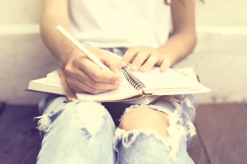 Eine Studentin sitzt auf dem Boden angelehnt an einer Wand, sie schreibt etwas in ein Notizbuch auf ihrem Schoß.